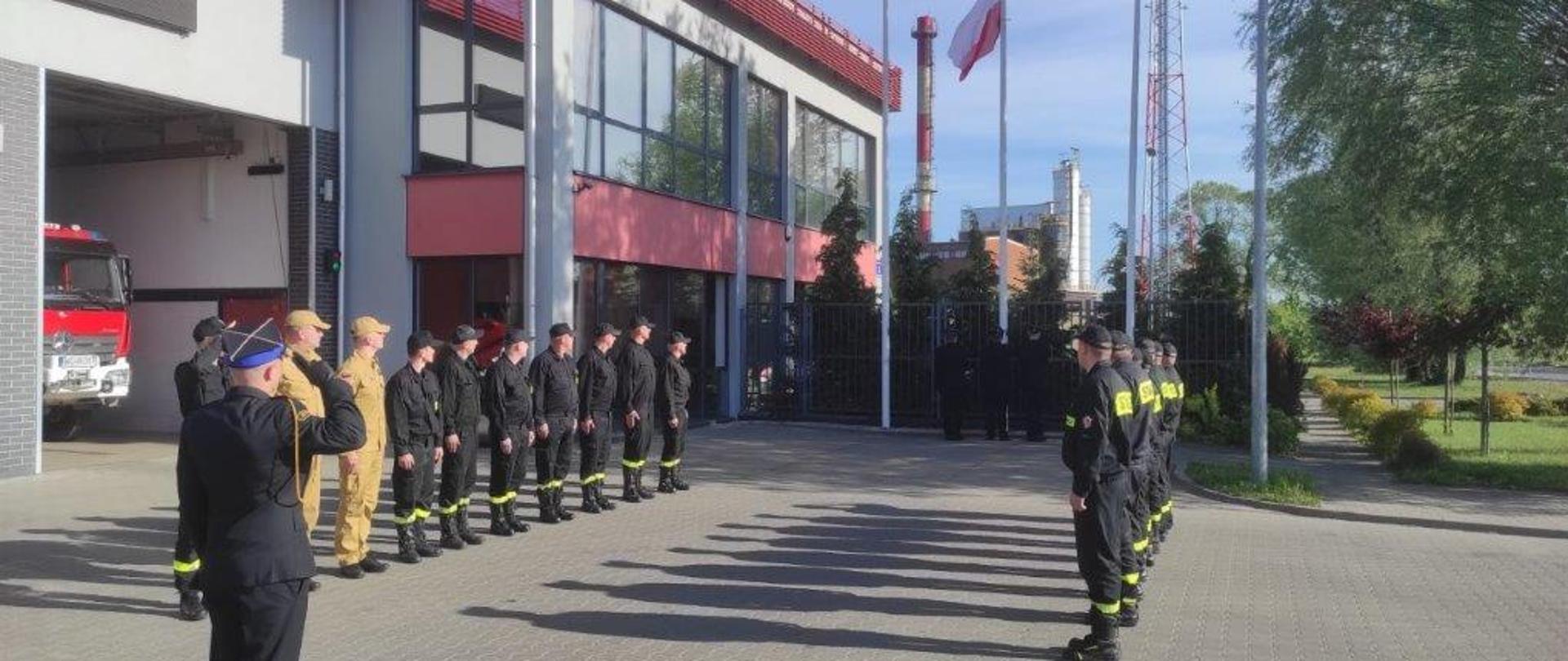 Zdjęcie przedstawia funkcjonariuszy Państwowej Straży Pożarnej. Funkcjonariusze ustawieni w dwóch szeregach zwróconych frontem ku sobie. Ubrani są w ubrania koszarowe w kolorze czarnym oraz żółtym piaskowym. Pomiędzy nimi stoi dowódca jednostki. Ubrany jest w galowy mundur. Strażacy ustawieni są w pozycji zasadniczej „na baczność” podczas podniesienia flagi państwowej na maszt. W oddali widać poczet flagowy, który dokonuje podniesienia flagi. Po lewej stronie dodatkowo widać otwartą bramę garażową. W garażu widać pojazd pożarniczy w kolorze czerwonym z białymi elementami zabudowy. Zdjęcie wykonane przed Jednostką Ratowniczo – Gaśniczą numer 2 w Radomiu. 