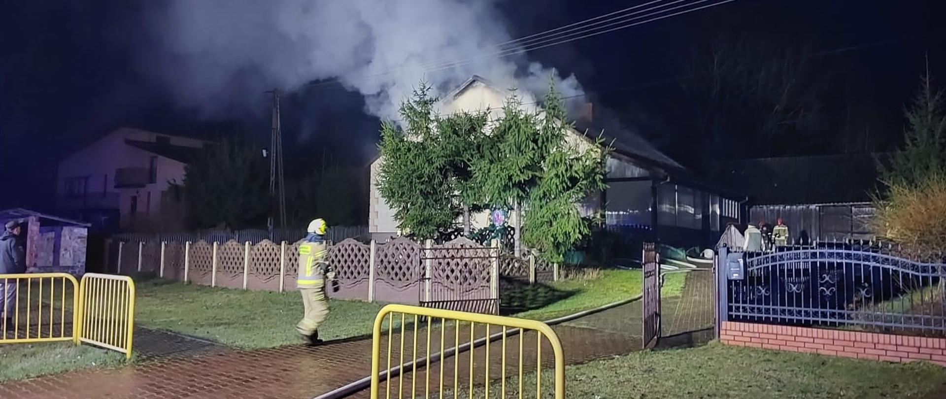 Zdjęcie przedstawia pożar budynku mieszkalnego w nocy. Z dachu i górnej części białego domu z ciemnym dachem wydobywa się gęsty, biały dym. Przed domem rosną trzy duże, zielone iglaste krzewy lub małe drzewa, które częściowo zasłaniają frontową ścianę. Na pierwszym planie, po lewej stronie, widać żółte, metalowe barierki i fragment chodnika. Na ścieżce przed domem stoi strażak w żółtym, odblaskowym ubraniu i hełmie, idący w kierunku budynku. W prawym dolnym rogu zdjęcia widać fragment ogrodzenia. W tle, po lewej stronie, widoczny jest inny budynek mieszkalny, a także słupy i linie energetyczne. Cała scena jest oświetlona jedynie światłem pochodzącym z pożaru i otoczenia, co podkreśla porę nocną i dramatyzm sytuacji.