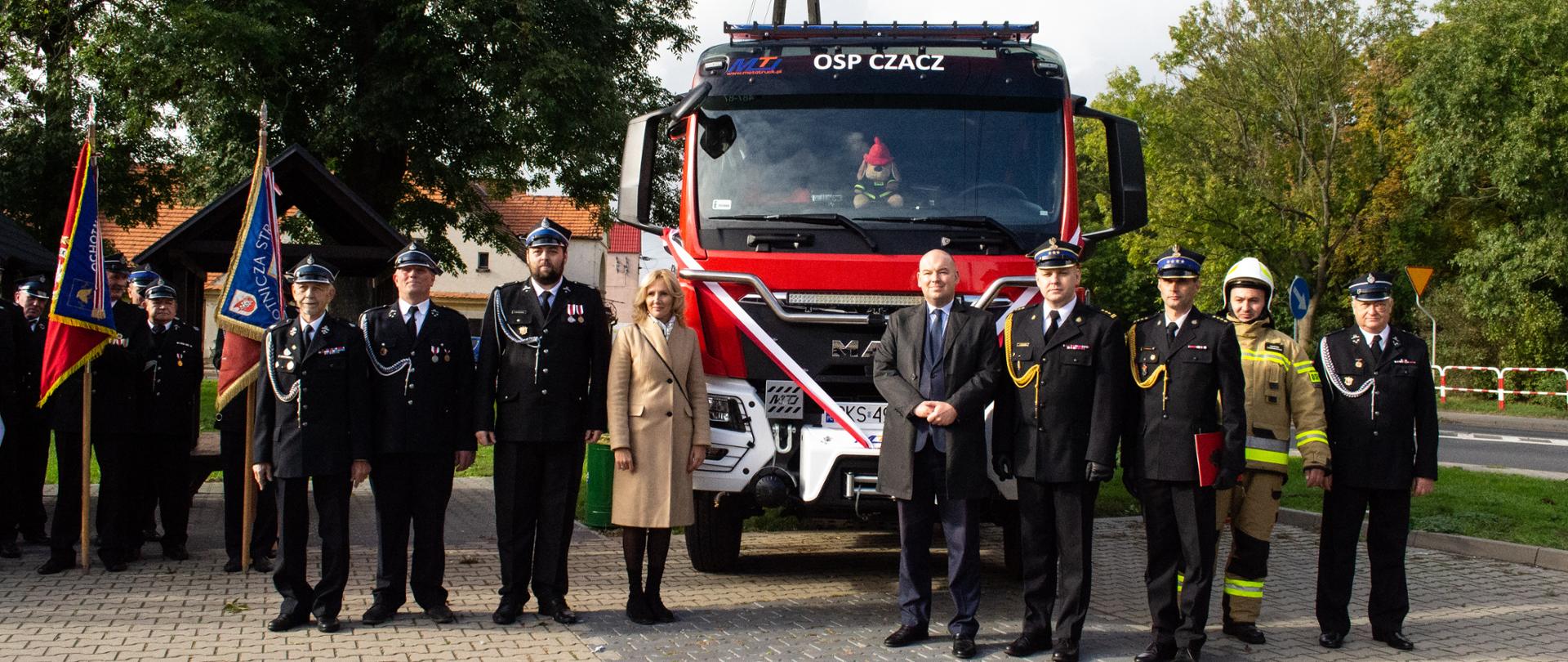 JUBILEUSZ 90-LECIA OSP CZACZ I PRZEKAZANIE NOWEGO SAMOCHODU. Na zdjęciu widać gości i strażaków przed samochodem strażackim, w tle drzewa.