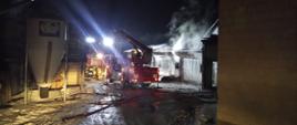 Rozstawiony podnośnik hydrauliczny w porze nocnej, wykorzystany podczas działań gaśniczych . Pożar budynku gospodarczego opanowany trwa dozorowanie miejsca pożaru. Miejsce zdarzenia jest oświetlone. Strażacy stoją przy samochodach ratowniczych obserwują ugaszony budynek .