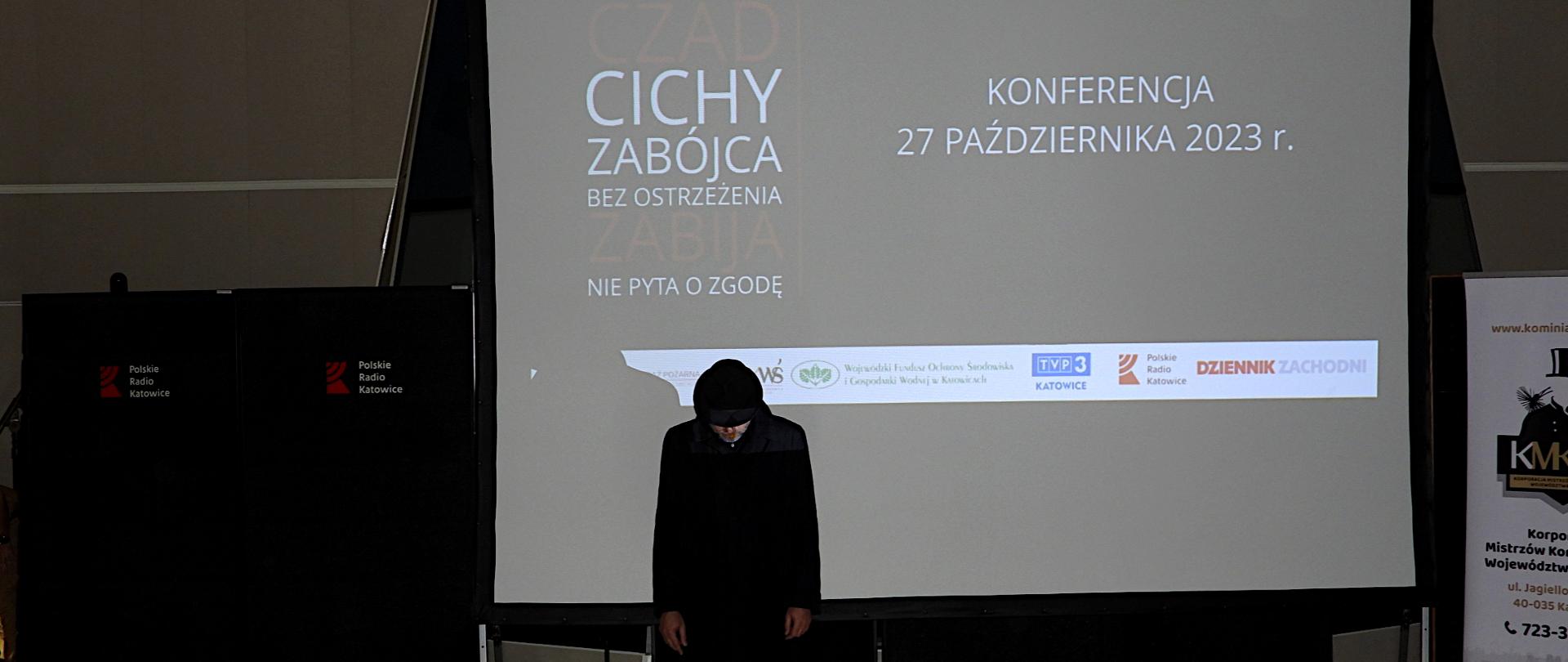 Na auli z wygaszonym światłem na wprost ekran, na którym wyświetlany jest napis na środku: Konferencja 27 października 2023 roku. Z lewej strony napis: Bezwonny, niewidoczny czad cichy zabójca bez ostrzeżenia zabija nie pyta o zgodę. Pod spodem logotypy partnerów i patronów akcji. Przed ekranem stoi mężczyzna ze spuszczoną głową. Ubrany w ciemny płaszcz i kapelusz.