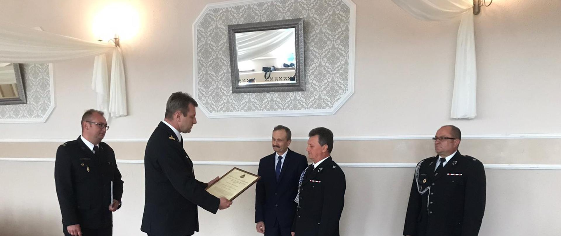 Zdjęcie przedstawia funkcjonariuszy Ochotniczej Straży Pożarnej oraz Państwowej Straży Pożarnej w mundurach wyjściowych podczas przekazywania decyzji Komendanta Głównego