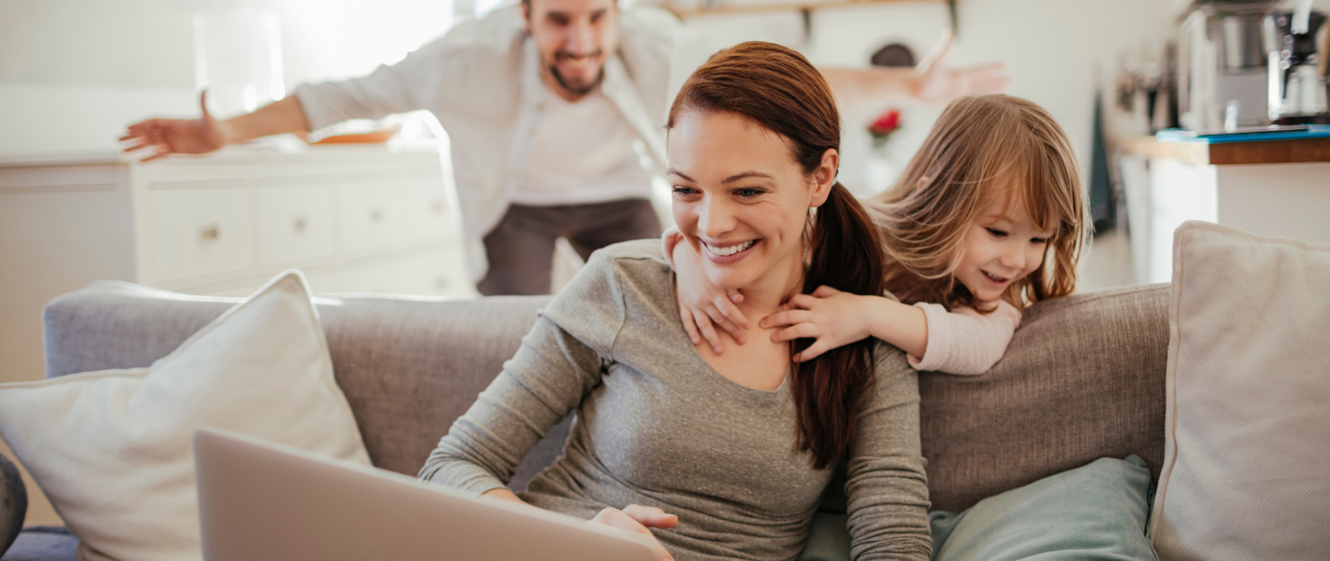 kobieta siedzi na kanapie, przed nią komputer, obok dziecko i mężczyzna. Rodzina w pomieszczeniu