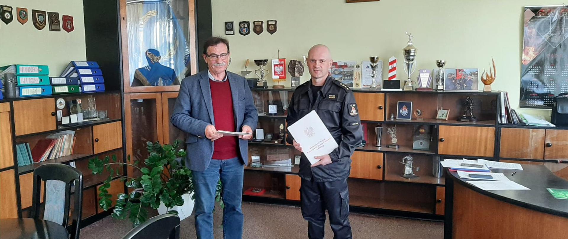 Komendant Miejski Państwowej Straży Pożarnej w Elblągu oraz Pana Konrada Murzyńskie Prezes Zarządu PRZEMBUD GDAŃSK. Panowie stoją w centralnej części gabinetu komendanta, trzymają w rękach teczki z podpisaną umową.