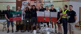 W dużej hali stoi grupa młodych ludzi, trzymają polskie flagi i zielono-czarno-czerwoną flagę z napisem ADH, przed nimi mały robot na czterokołowym podwoziu.