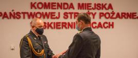 Zdjęcie przedstawia dwóch strażaków komendy miejskiej w mundurach wyjściowych koloru czarnego na tle czerwonego napisu Komenda Miejska Państwowej Straży Pożarnej w Skierniewicach. Jeden ze strażaków wręcza drugiemu rozkaz personalny.