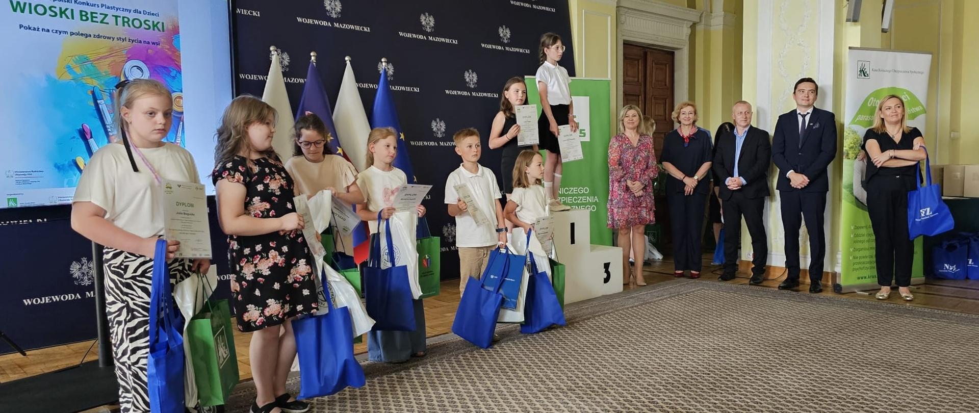 W reprezentacyjnej sali na tle rollupów z logo KRUS pozuje do zdjęcia siedem dziewczynek i jeden chłopiec. Dzieci trzymają w dłoniach dyplomy i niebieskie i zielone torby z nagrodami.