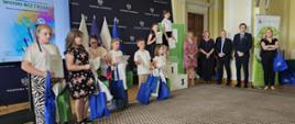 W reprezentacyjnej sali na tle rollupów z logo KRUS pozuje do zdjęcia siedem dziewczynek i jeden chłopiec. Dzieci trzymają w dłoniach dyplomy i niebieskie i zielone torby z nagrodami.

