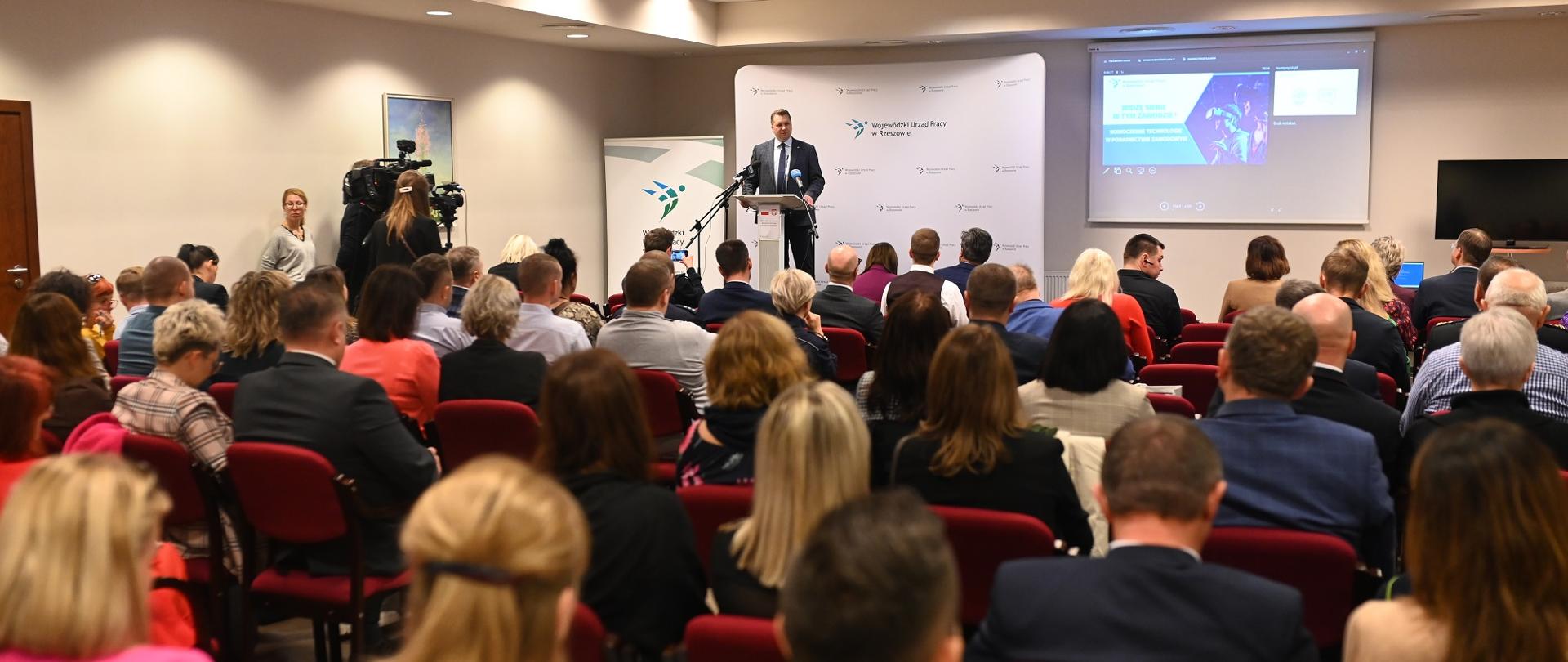 Widok z tyłu na salę, na krzesłach siedzą ludzie, przed nimi ktoś przy mównicy mówi do mikrofonu.