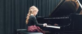 Dziewczynka gra na fortepianie, w tle czarna kurtyna.
