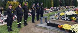 Widok z przodu. Od lewej trzech strażaków zmiany służbowej w ubraniach koszarowych, kolejno funkcjonariusze Komendy Powiatowej w umundurowaniu galowym. Strażacy stoją w szeregu salutując przed nagrobkiem. 