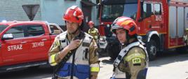 Na pierwszym planie dwóch strażaków w ubraniach bojowych, biało-niebieskich kamizelkach i czerwonych hełmach. W tle widoczne dwa czerwone wozy bojowe. 