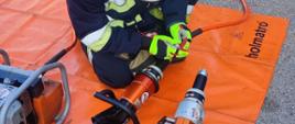 Na zdjęciu widać strażaka, który sprawia sprzęt hydrauliczny. Strażak podłącza przewód hydrauliczny do narzędzia - nożyc hydraulicznych.