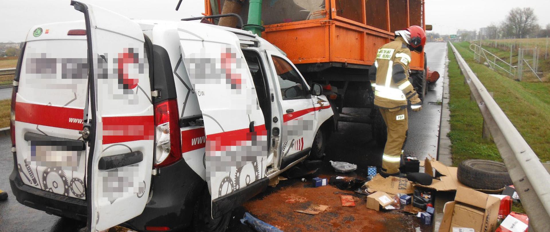 Na zdjęciu widać rozbity samochód osobowy z pootwieranymi drzwiami i tył przyczepy ciągnika. Na jezdni plama substancji ropopochodnej, która wyciekła z uszkodzonej osobówki oraz porozrzucane przedmioty. Na poboczu leży urwane koło.