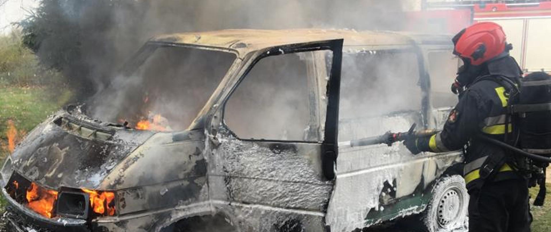 Zdjęcie przedstawia samochód typu bus w płomieniach, strażak ubrany u sprzęt ochrony dróg oddechowych gasi go pianą.
