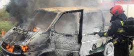 Zdjęcie przedstawia samochód typu bus w płomieniach, strażak ubrany u sprzęt ochrony dróg oddechowych gasi go pianą.