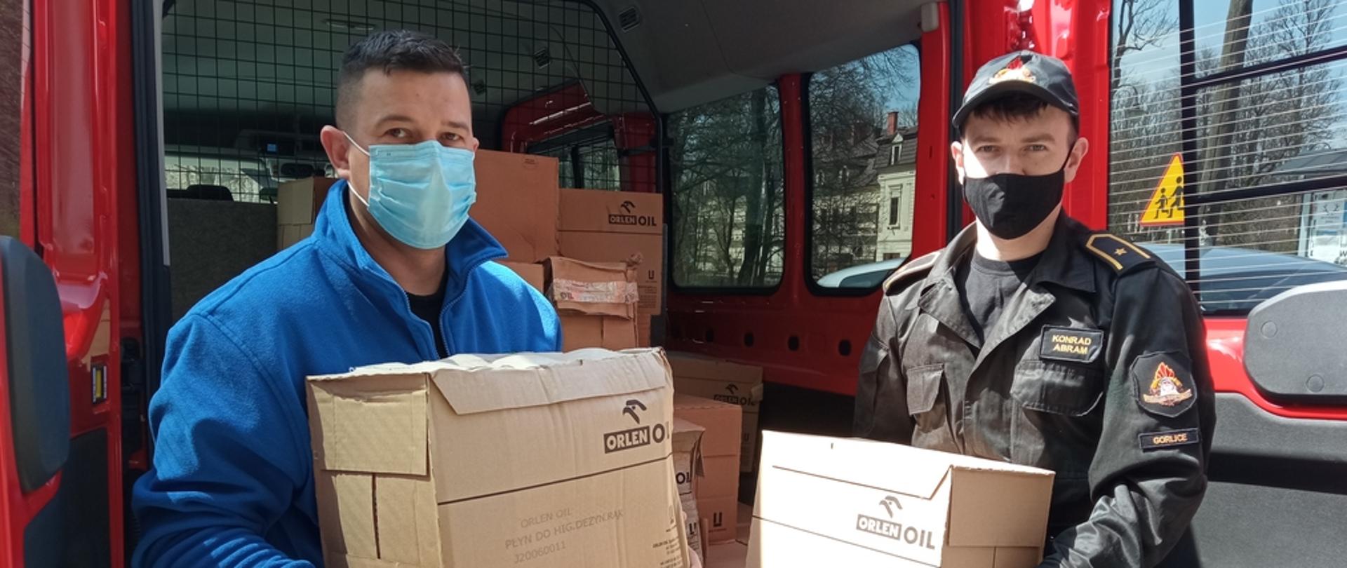 Na zdjęciu widoczni strażacy za placami samochód strażacki z kartonami z płynem do dezynfekcji i maseczkami. Po lewej strażak z OSP w ubraniu cywilnym w polarze niebieskim i niebieskich dżinsach po prawej w czarnym ubraniu dowódczo sztabowym w czapce z daszkiem strażak z PSP. Strażacy trzymają w rękach pudełka kartonowe z płynem do dezynfekcji.