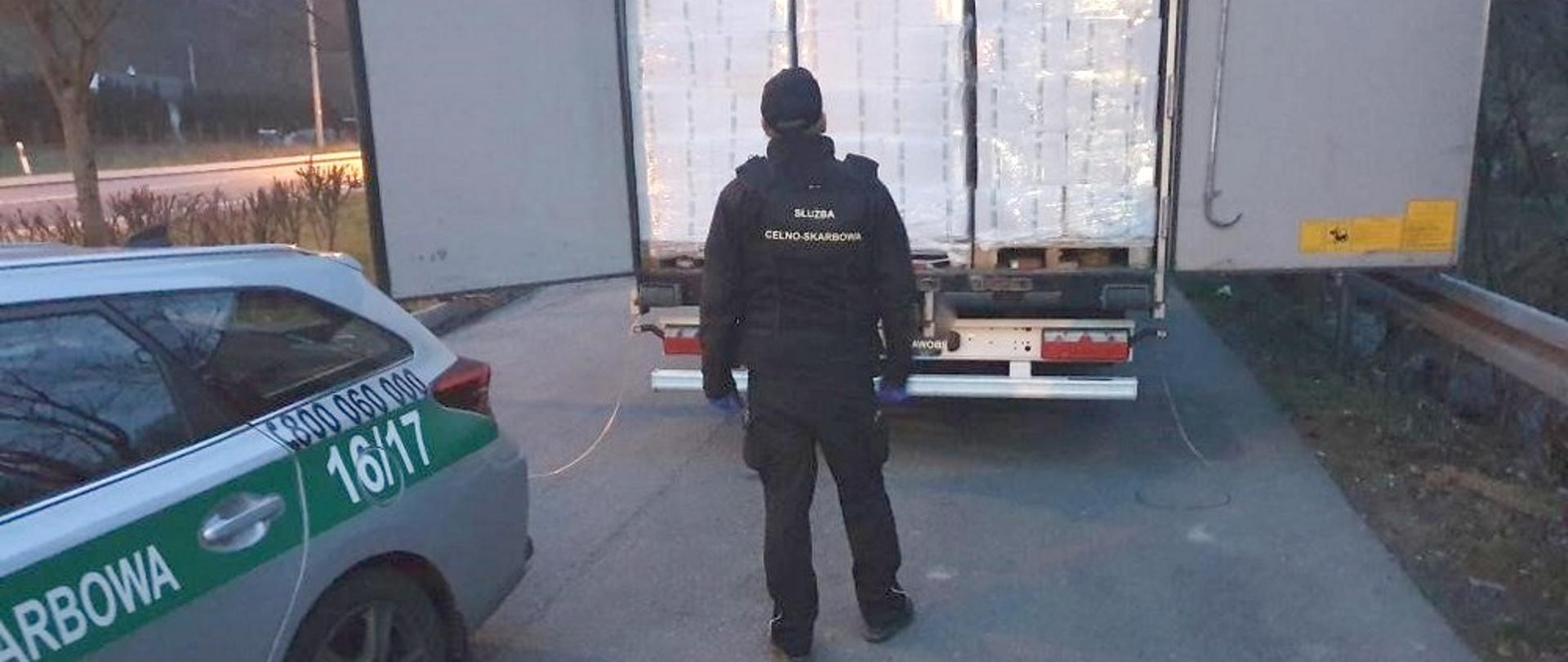 Funkcjonariusz KAS z tyłu samochodu ciężarowego z otwarta przestrzenią ładunkową, z lewej strony fragment radiowozu Służby Celno-Skarbowej.