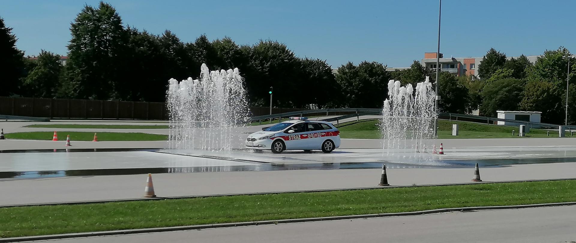 na zdjęciu strażacki samochód osobowy jadący po torze do symulacji poślizgów, widać2 bariery wodne, jezdnie płyty poślizgowej w tle drzewa