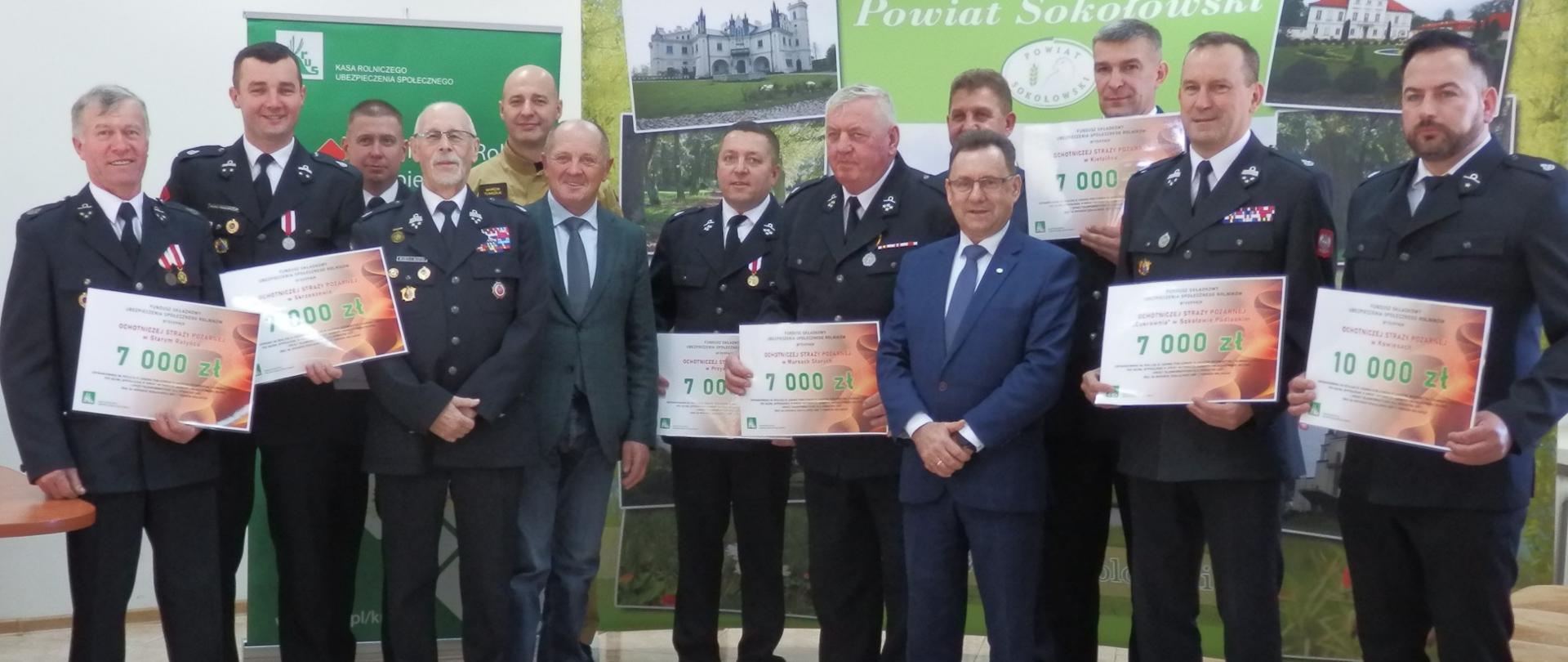 Trzynastu mężczyzn stoi tle banerów KRUS i Powiatu Sokołowskiego, część z nich w galowych mundurach strażaka, trzymają plansze z kwotą otrzymanego wsparcia.