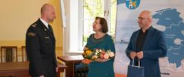 Starosta Lęborski Pani Alicja Zajączkowska wraz z członkiem Zarządu Powiatu składają gratulacje brygadierowi Leszkowi Formela z okazji powołania na stanowisko Komendanta Powiatowego Państwowej Straży Pożarnej w Lęborku. Starosta trzyma w rękach bukiet kwiatów obok niej członek zarządu trzyma niebieską torbę okolicznościową.