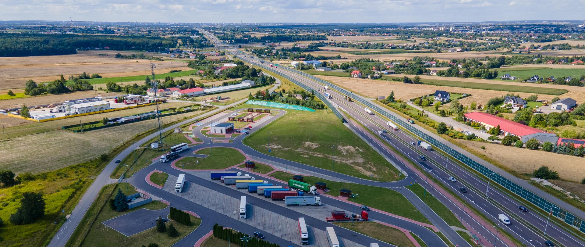 Obraz przedstawia widok na MOP przy drodze S12.Po prawej stronie znajduje się asfaltowa nawierzchnia drogi S12, po lewej budynki sanitariatu, parking oraz wiaty piknikowe i plac zabaw. Na parkingu stoi sporo samochodów, głównie ciężarowych.