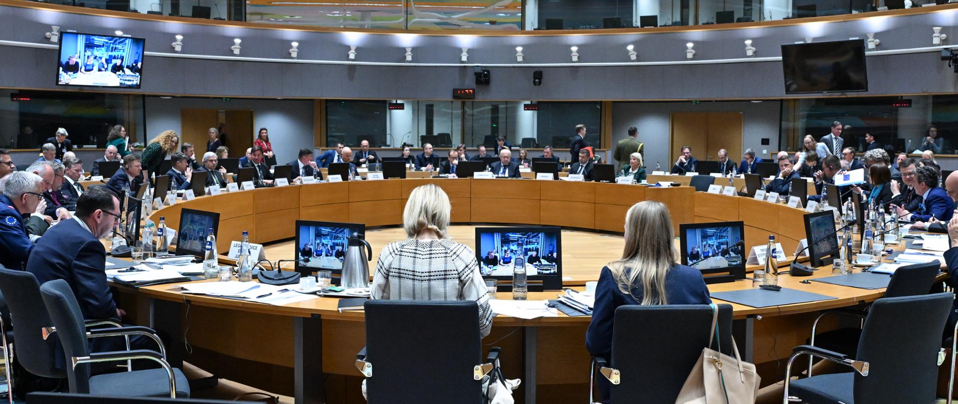 An international meeting is taking place in a large, modern conference room with a circular table arrangement. Participants in formal attire are seated around the tables, with monitors, documents, and microphones placed in front of them. The colorful ceiling and screens throughout the room give the interior a modern appearance.
