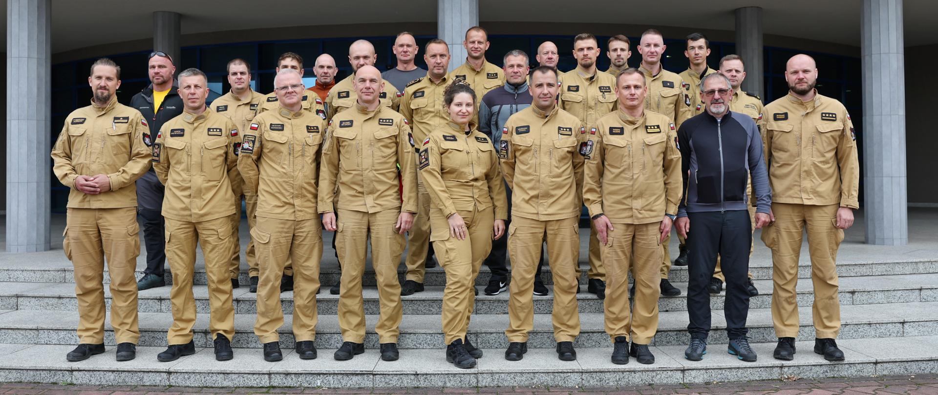 Grupowe zdjęcie uczestników szkolenia ratowniczego stojących na schodach przed budynkiem. Większość osób ubrana jest w piaskowe mundury strażackie, pozostali w ciemną odzież.
