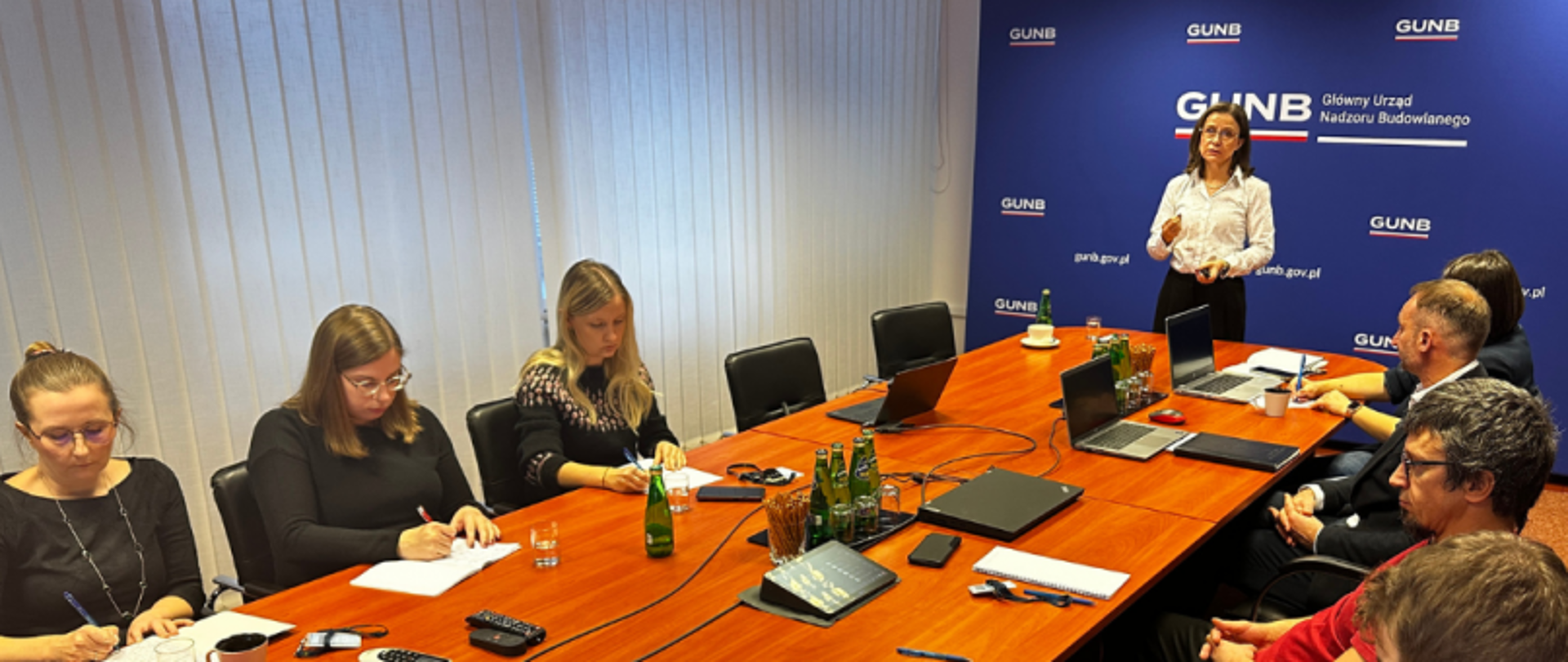 Grupa osób siedząca przy dużym stole konferencyjnym, jedna osoba stoi i przemawia; na ścianie napis „GUNB – Główny Urząd Nadzoru Budowlanego” oraz adres „gunb.gov.pl”.