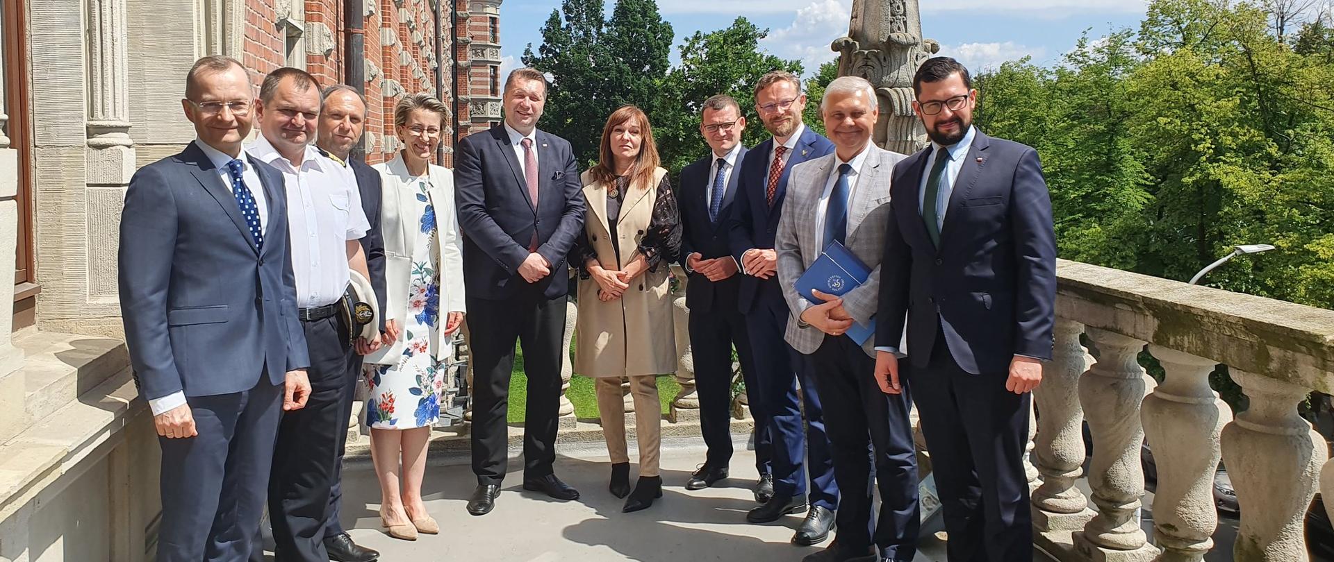 Minister w otoczeniu rektorów stoi na balkonie, za nimi zieleń i niebo.