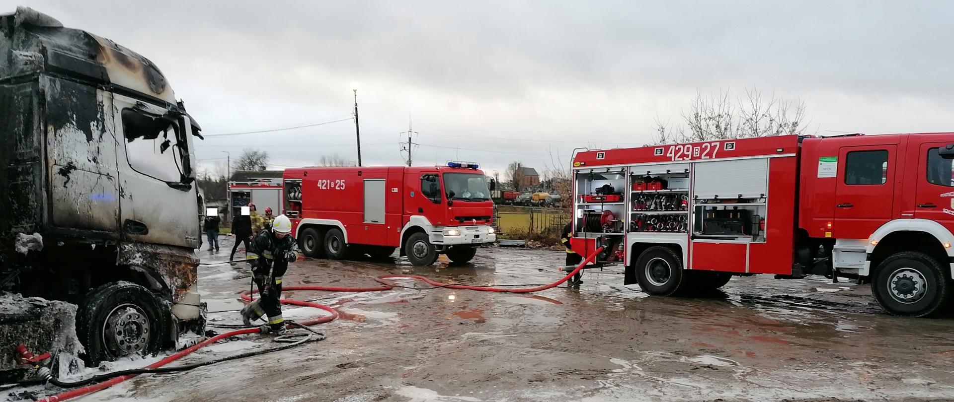 Z lewej spalona kabina samochodu ciężarowego. Przy niej strażak OSP w aparacie powietrznym. W tle samochody straży pożarnej.