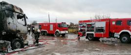 Z lewej spalona kabina samochodu ciężarowego. Przy niej strażak OSP w aparacie powietrznym. W tle samochody straży pożarnej.