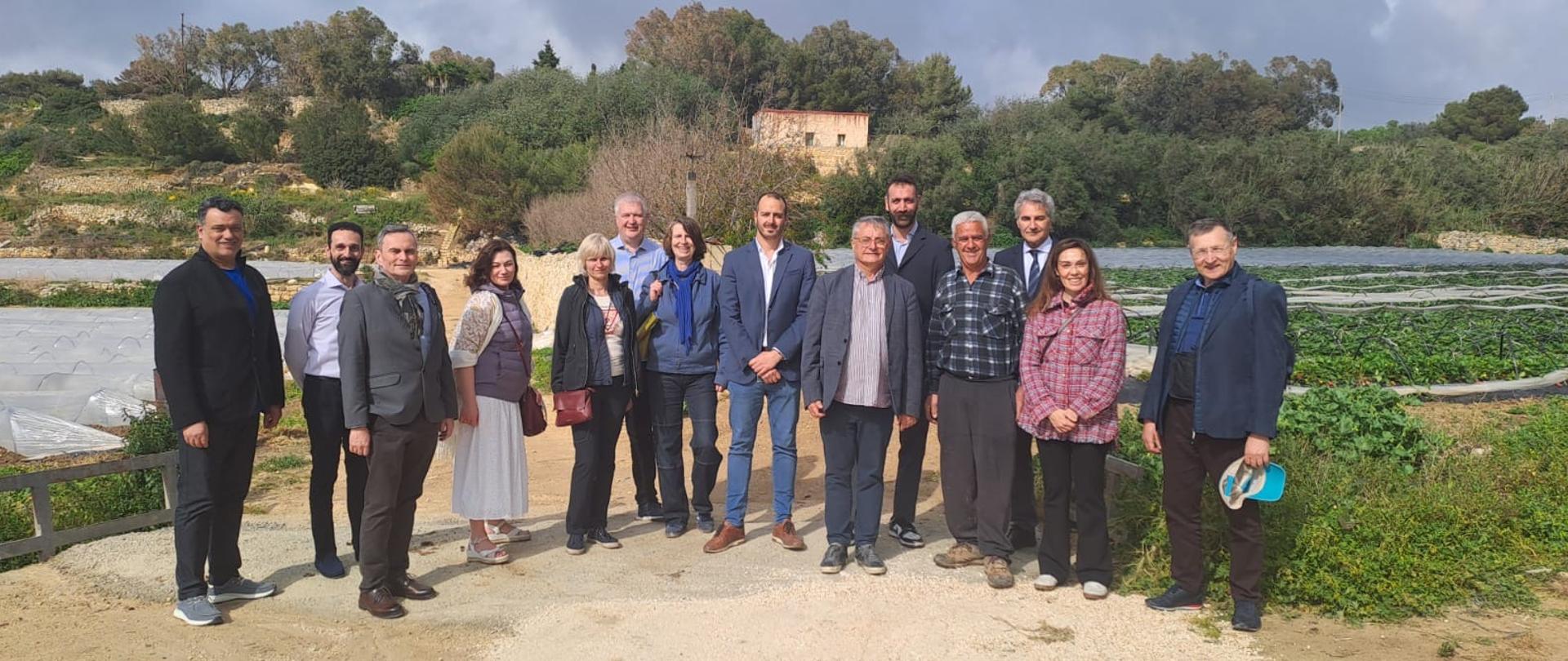 Kilka osób pozuje do pamiątkowego zdjęcia w gospodarstwie. Na drugim planie uprawa pod folią.