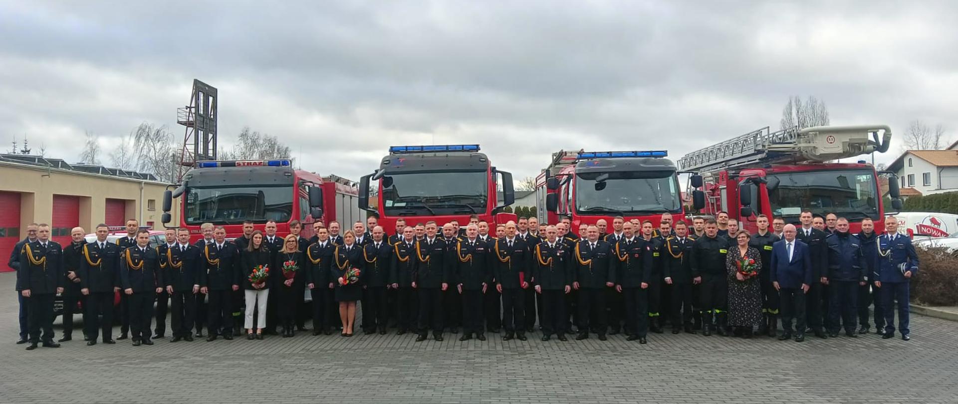 Zdjęcie przedstawia strażaków i zaproszonych gości po uroszystej zbiórce, na tle czterech samochodów pożarniczych.