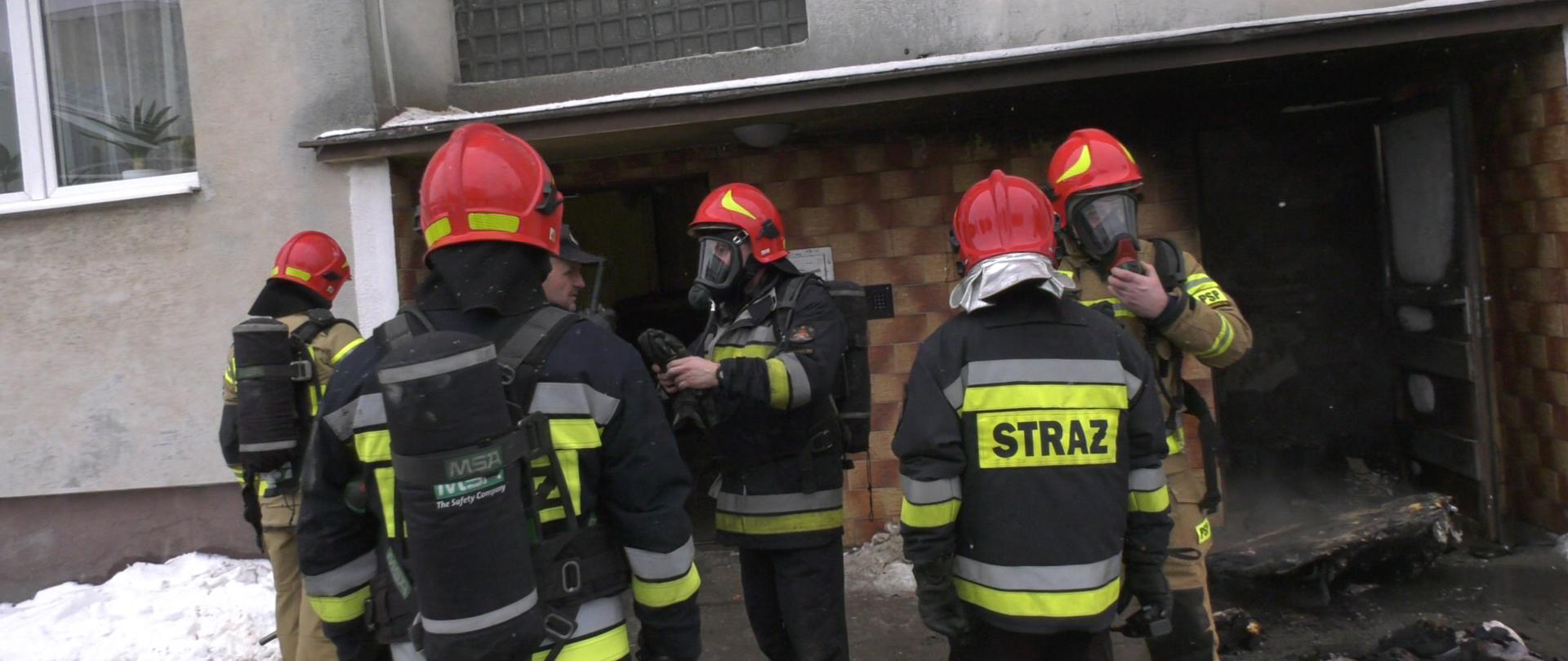 Na pierwszym planie strażacy ubierający się w aparaty powietrzne przed klatką budynku wielorodzinnego 