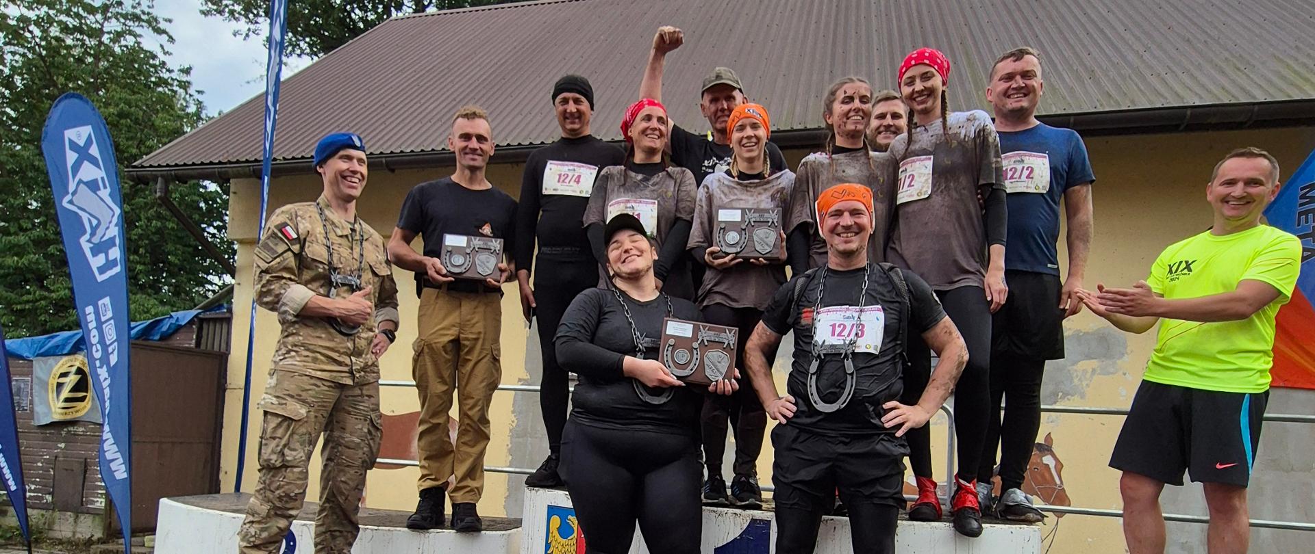 Grupa uśmiechniętych zawodników stoi na pierwszym stopniu podium, a obok nich przy podium stoi żołnierz w mundurze polowym i mężczyzna w żółtej koszulce. W tle brązowy dach budynku wysokie drzewa i niebo. 