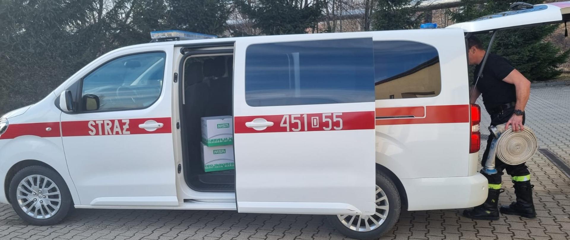 Funkcjonariusz straży pożarnej w czarnych ubraniu koszarowy uzupełnia sprzęt przekazywany w darze pomocy dla Ukrainy. Na zdjęciu widoczne węże strażackie oraz hełmy. znajdujące się w ustawionym centralnie samochodzie operacyjnym bus. Pojazd koloru białego z widocznym oznaczeniem operacyjnym 451D55. Samochód ustawiony na placu Komendy Powiatowej Państwowej Straży Pożarnej w Kamiennej Górze. w Kamiennej Górze.