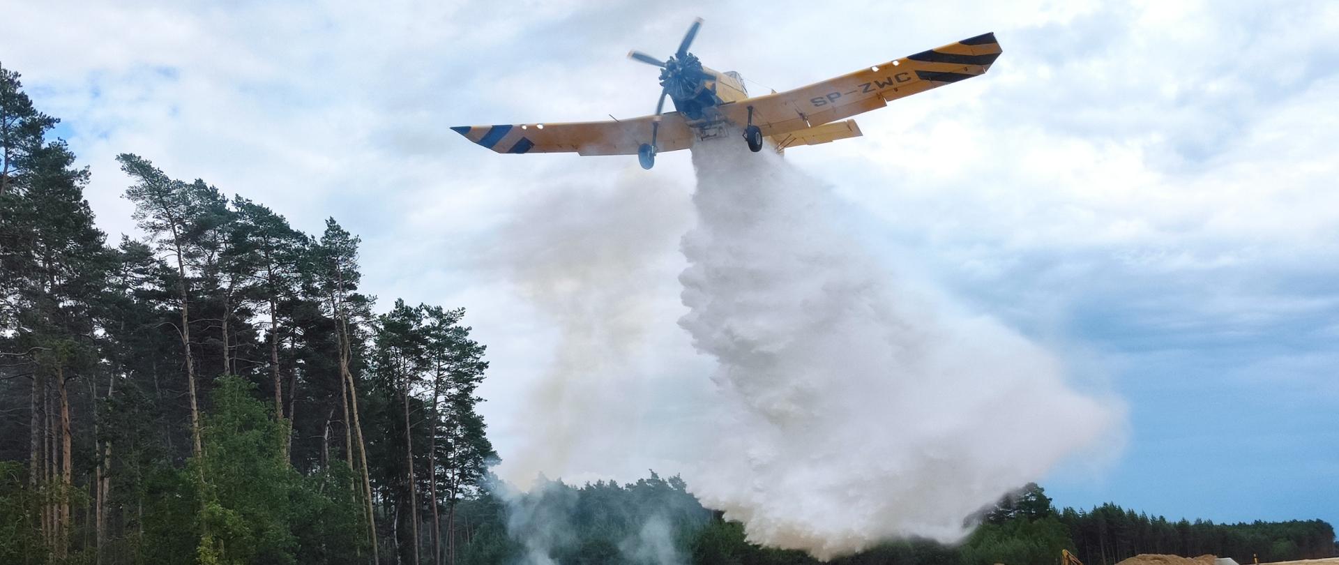 Na zdjęciu samolot gaśniczy wykonuje zrzut wody na ognisko, w tle widoczny las.