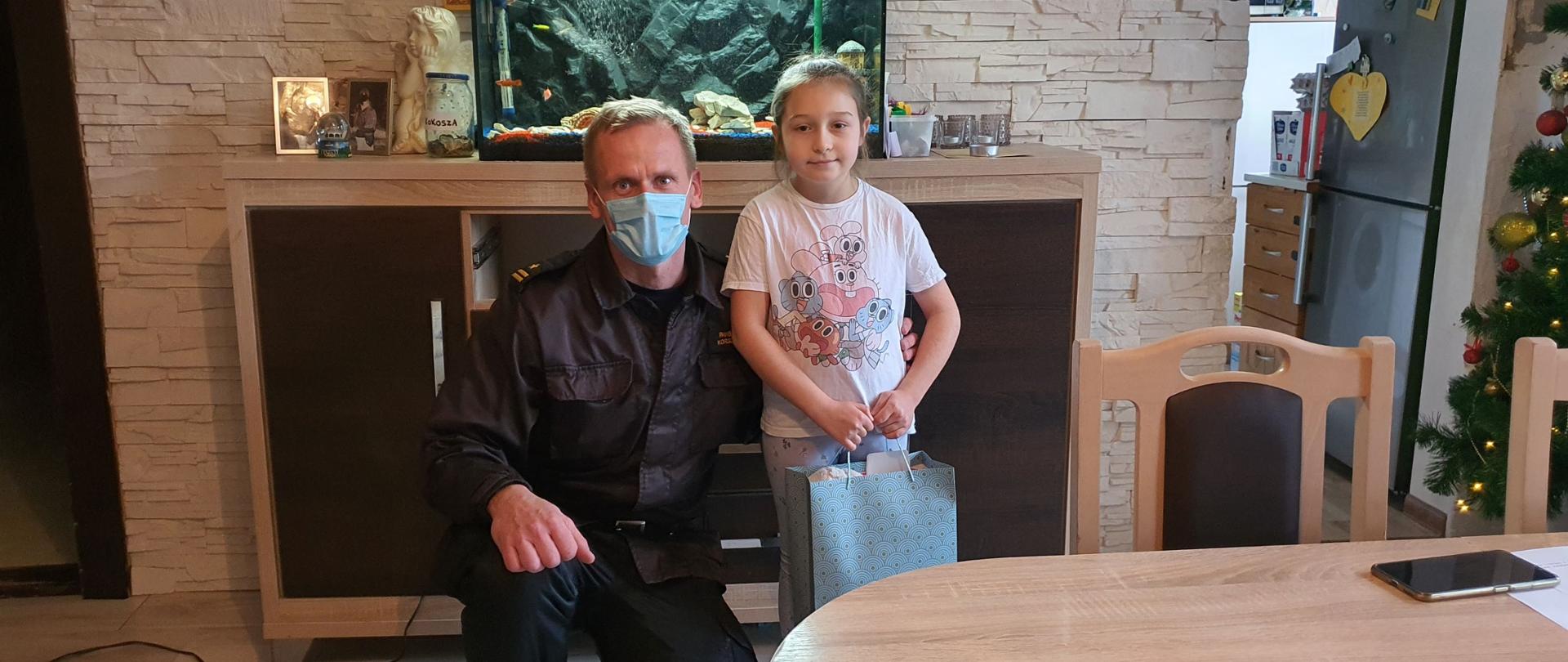 Zrobione wewnątrz pomieszczenia mieszkalnego zdjęcie przedstawia mężczyznę ubranego na czarno, który kucając pozuje do zdjęcia z małą dziewczynką. W tle komoda, na której stoi podświetlone akwarium.