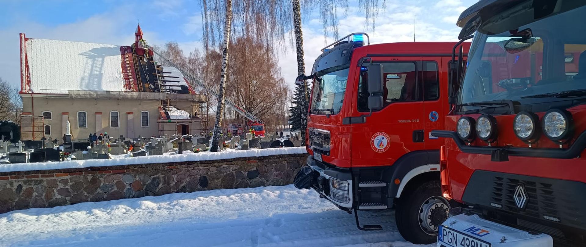 Zdjęcie zrobione z parkingu kościoła. Na zdjęciu widać ogrodzenie za którym jest cmentarz a w tle kościół na dachu którego powstał pożar. Po prawej stronie zdjęcia widać dwa samochody pożarnicze. W tle przy kościele widać rozłożoną autodrabinę, która jest wykorzystywana podczas działań. Jest pogodny zimowy dzień, widać leżący śnieg.