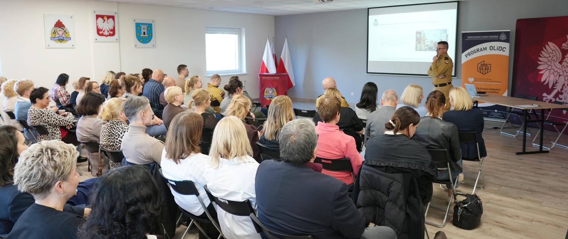 Szeroki kadr przedstawiający salę konferencyjną pełną słuchaczy, którzy uczestniczą w prelekcji prowadzonej przez umundurowanego strażaka