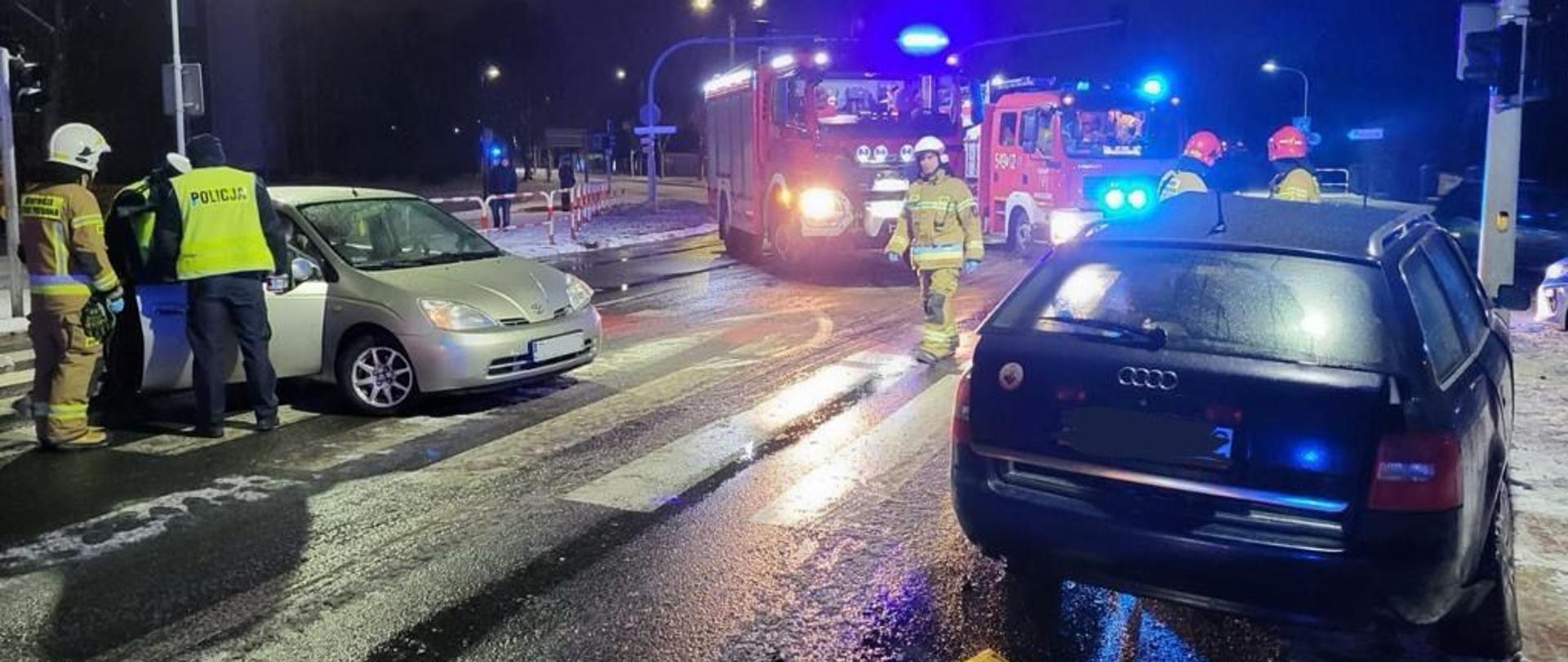 Samochody osobowe po zderzeniu znajdują się na drodze. W okolicy znajdują się pojazdy służb ratowniczych oraz ratownicy. Zdjęcie wykonane w porze nocnej.