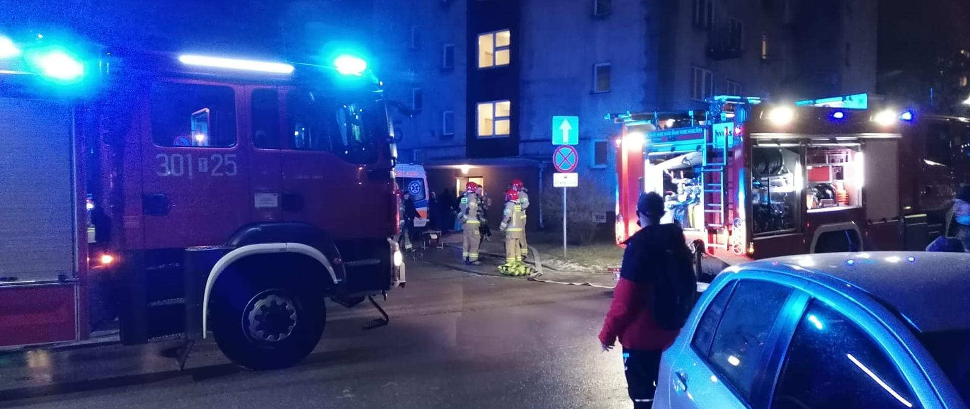 Na zdjęciu widzimy samochód pożarniczy biorący udział w akcji, na dalszym planie trzech ratowników przygotowanych do wejścia do budynku gdzie wybuchł pożar. W tle budynek wielorodzinny, w którym wybuchł pożar.