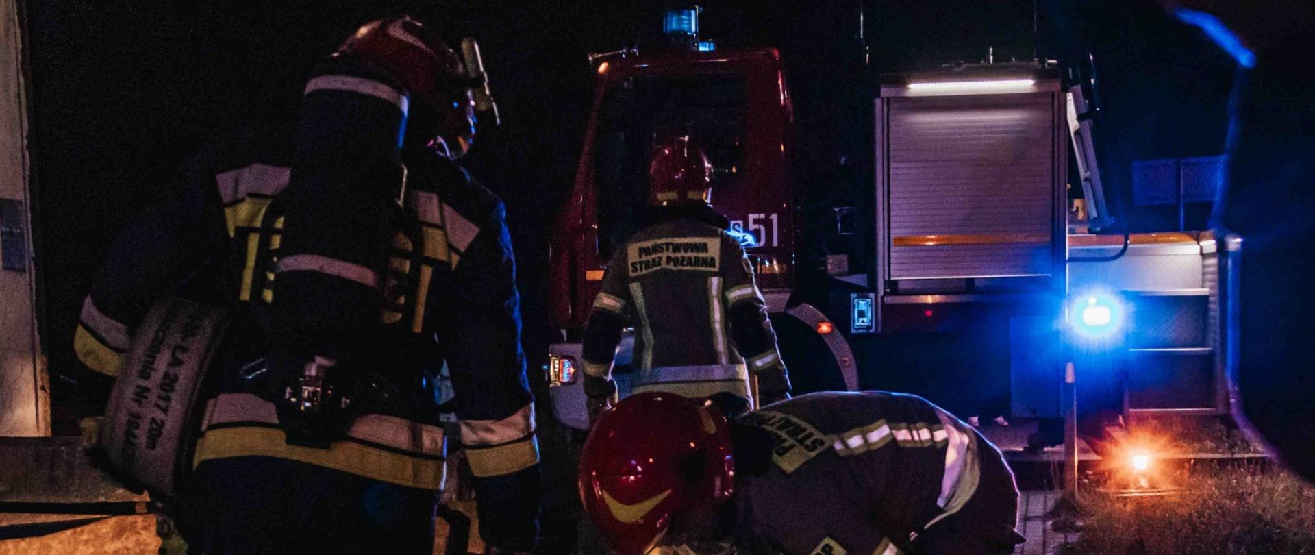 Nocna akcja strażaków przy oświetlonych wozach strażackich. Strażacy w hełmach i odblaskowych mundurach Państwowej Straży Pożarnej pracują przy sprzęcie, a niebieskie światła pojazdów rozświetlają otoczenie.