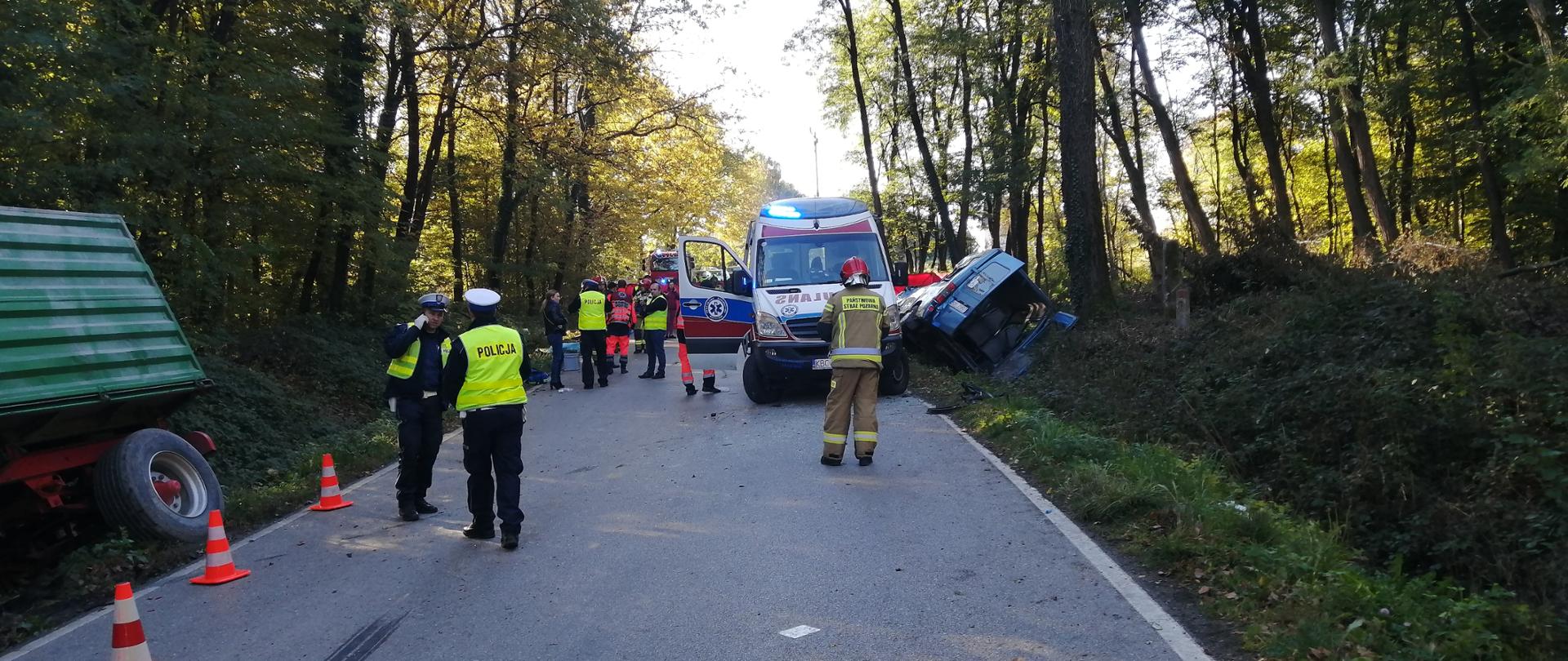 Zdjęcie w centralnej części przedstawia dwóch policjantów i strażaka, którzy stoją na drodze przed samochodem zespołu ratownictwa medycznego. Po prawej stronie zdjęcia znajduje się przewrócony bus, którym podróżowały osoby. Z lewej strony zdjęcia widać część przyczepu od ciągnika rolniczego