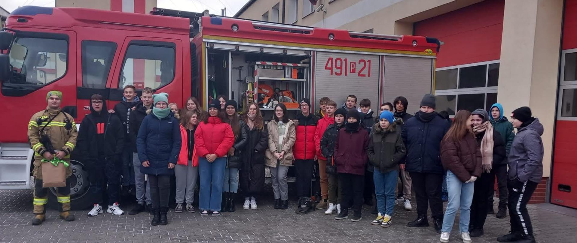 Zdjęcie grupowe uczniów odwiedzających krotoszyńską strażnicę ze strażakami na tle wozu bojowego. Uczniowie wizytowali u strażaków celem poznania specyfiki służby