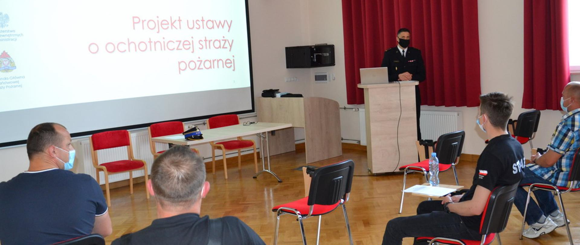 W sali na krzesłach siedzą druhowie OSP. Komendant w umundurowaniu galowym stoi za mównicą . na dużym ekranie prezentowany projekt ustawy o osp