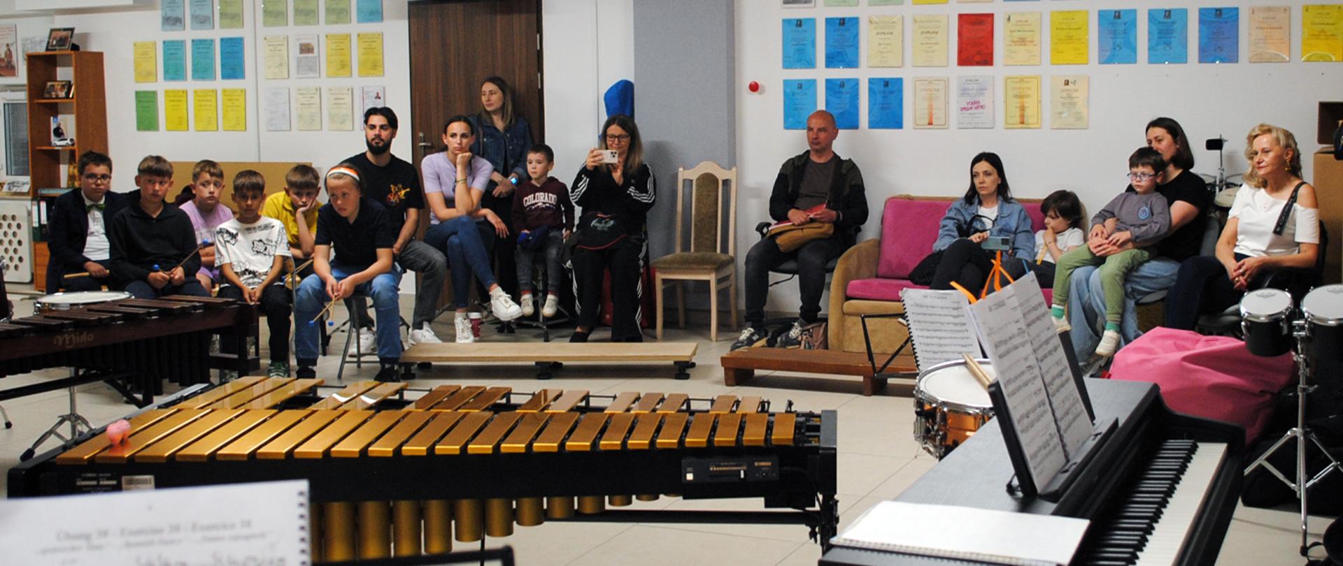 Grupa kilkunastu osób, dorosłych i dzieci siedzi w sali perkusji, przed nimi stoją instrumenty perkusyjne.