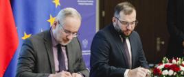 Minister funduszy i polityki regionalnej Grzegorz Puda i minister zdrowia Adam Niedzielski podpisują porozumienie w sprawie realizacji wybranych zadań z programu Fundusze Europejskie na Infrastrukturę, Klimat, Środowisko 2021–2027, ministrowie siedzą przy stole i podpisują dokumenty, za ich plecami flagi Polski i UE
