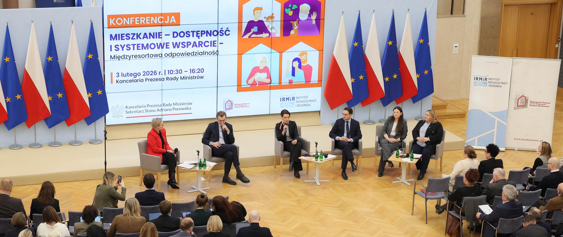Szeroki kadr, zdjęcie zrobione z góry. Na zdjęciu uczestnicy konferencji podczas Panelu Ministrów. W tle na środku widać ekran, na którym jest przedstawiona grafika z tytułem: Konferencja „MIESZKANIE – dostępność i systemowe wsparcie – międzyresortowa odpowiedzialność”, 3 lutego 2026 r., 10:30 – 16:20, Kancelaria Prezesa Rady Ministrów. Po obu stronach ekranu widać flagi Polski i Unii Europejskiej. Przed ekranem znajdują się paneliści Panelu Ministrów - od lewej: Moderatorka - Adriana Porowska, Sekretarz Stanu w Kancelarii Prezesa Rady Ministrów, Wiceprzewodnicząca Komitetu do spraw Pożytku Publicznego, Jan Szyszko, Sekretarz Stanu w Ministerstwie Funduszy i Polityki Regionalnej, Marzena Okła-Drewnowicz, Sekretarz Stanu w KPRM, Tomasz Lewandowski, Podsekretarz Stanu w Ministerstwie Rozwoju i Technologii, Katarzyna Nowakowska, Podsekretarz Stanu w Ministerstwie Rodziny, Pracy i Polityki Społecznej, Maja Nowak, Sekretarz Stanu w Ministerstwie Rodziny, Pracy i Polityki Społecznej, Pełnomocnik ds. Osób z Niepełnosprawnościami. Lewą, jak i prawą stronę sali zajmują uczestnicy wydarzenia.